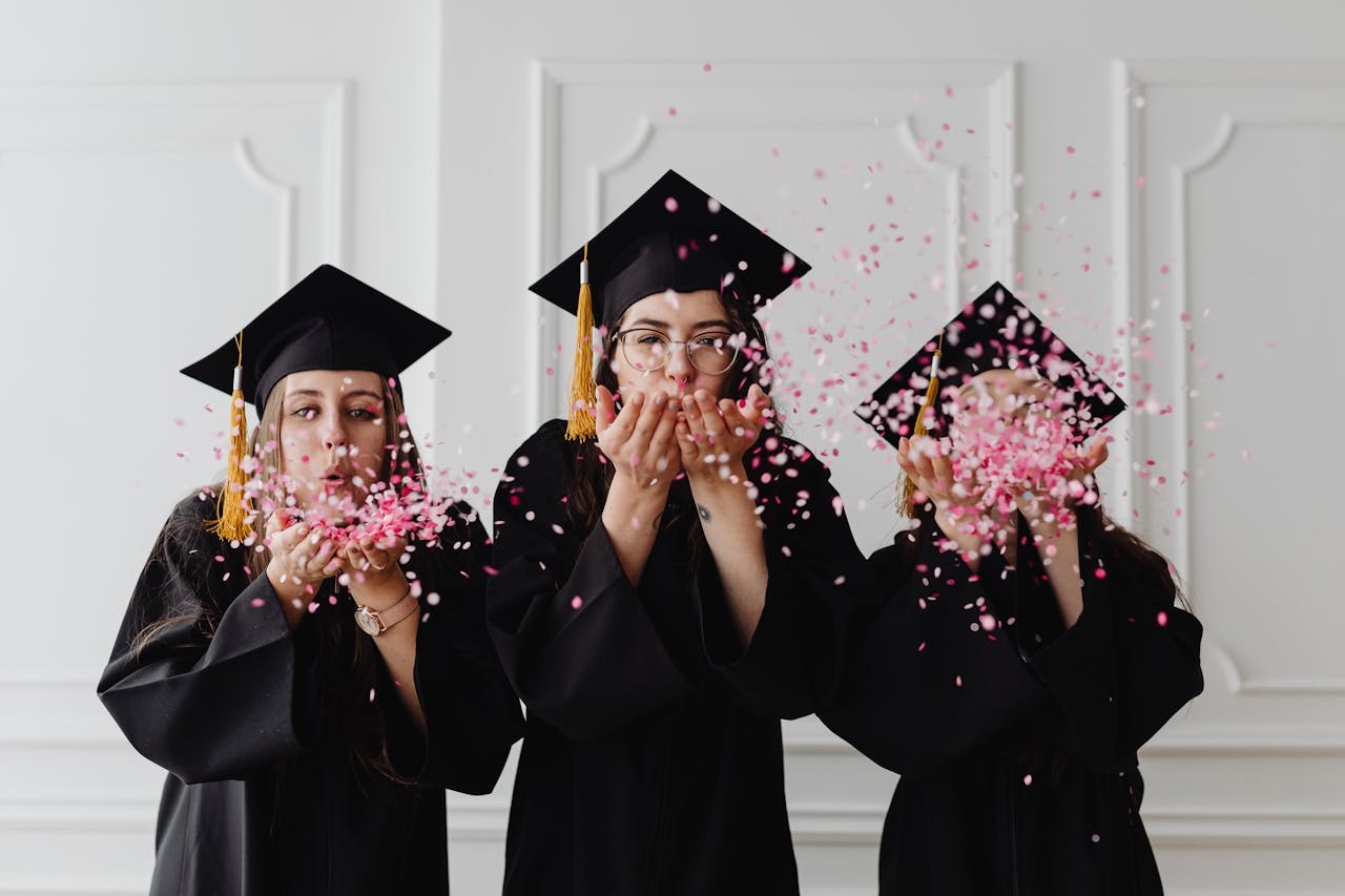 Three women in graduation gowns celebrate by blowing confetti indoors.